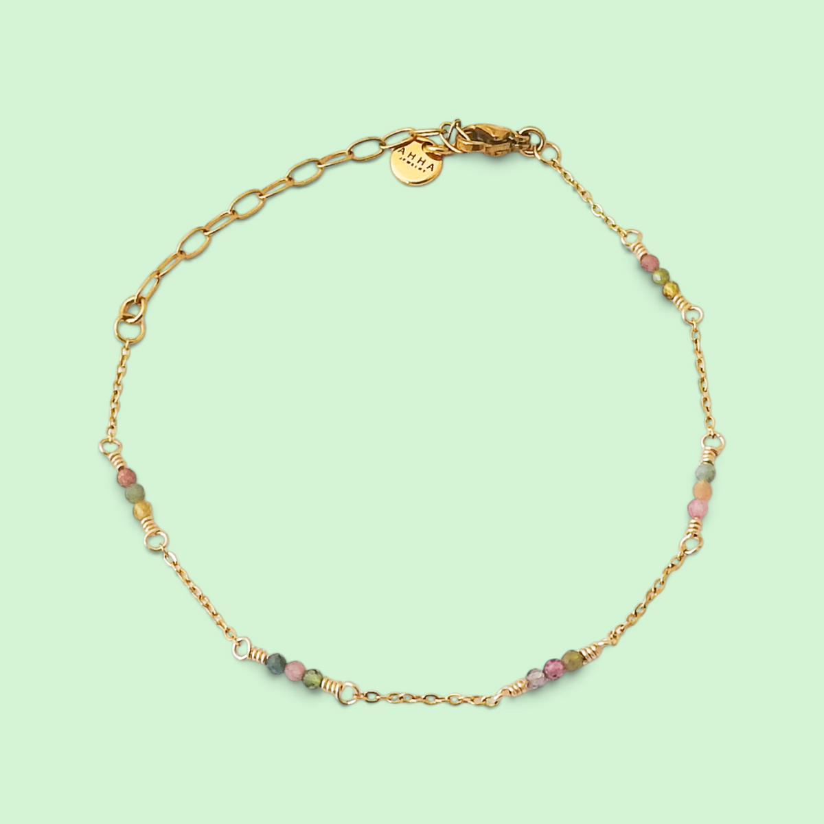A delicate gold chain bracelet with small multicolored beads and gold spacer rings, displayed on a light mint green background. A round gold tag with the brand name 'L'ENFANT' is visible near the clasp.