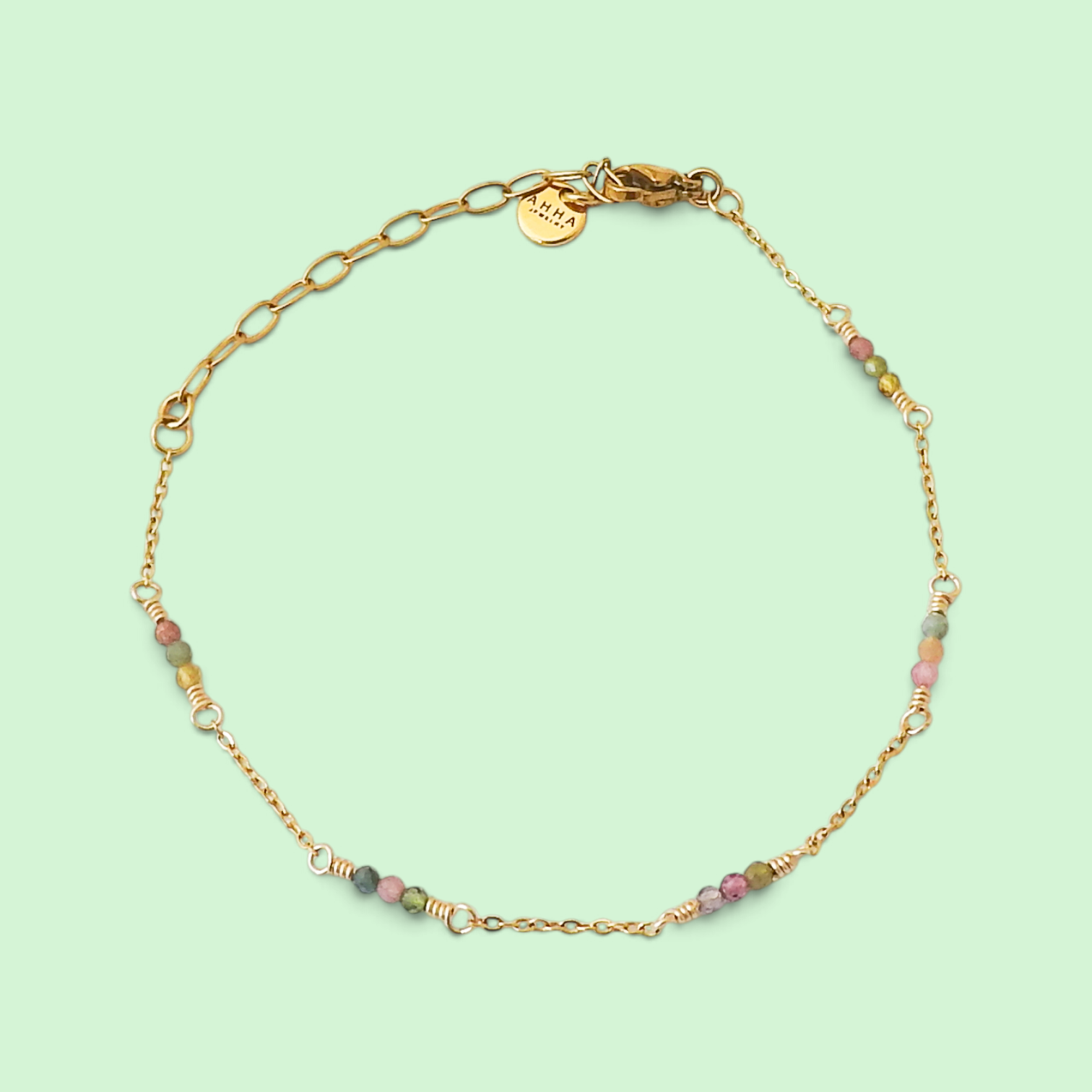 A delicate gold chain bracelet with small multicolored beads and gold spacer rings, displayed on a light mint green background. A round gold tag with the brand name 'L'ENFANT' is visible near the clasp.