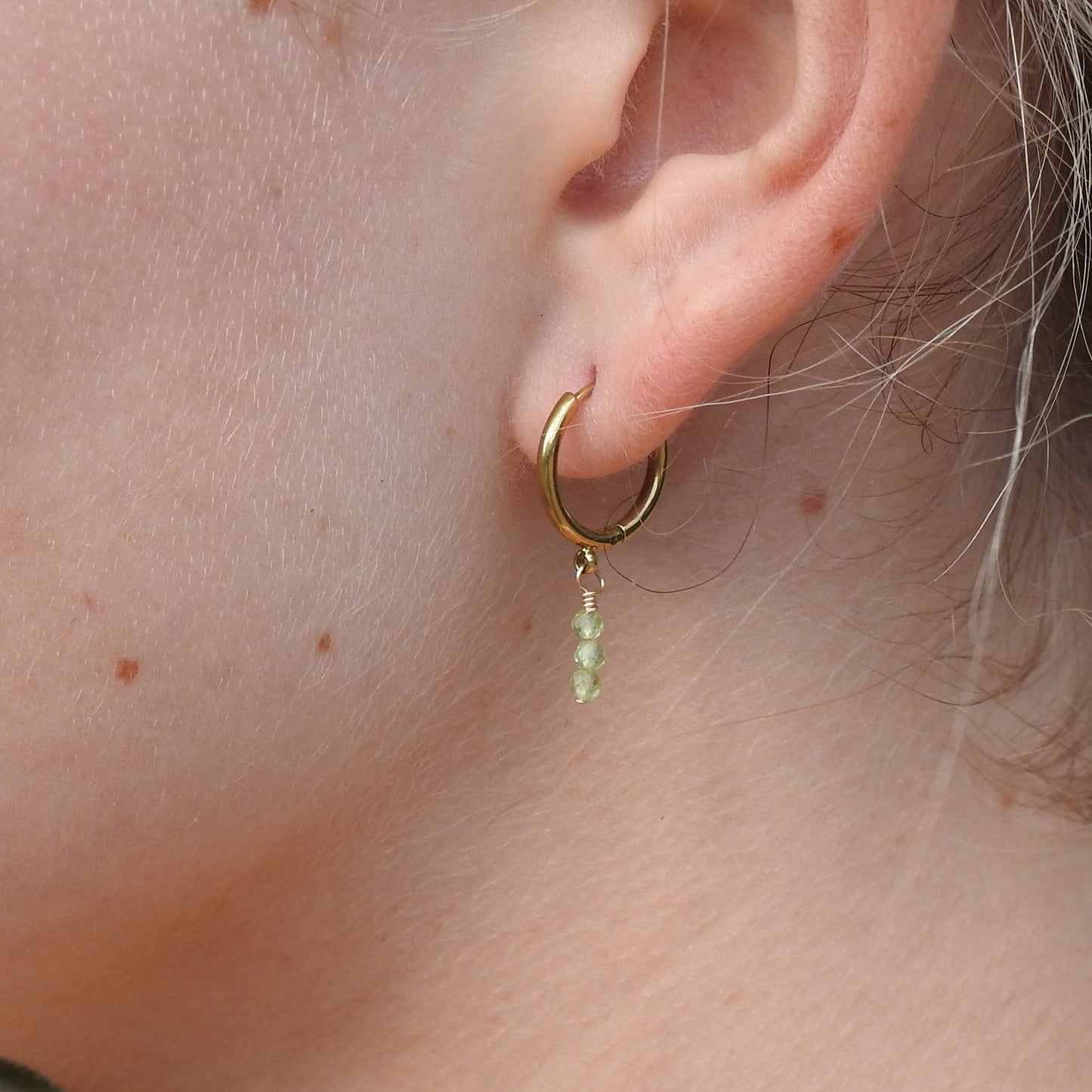 A close-up of a person's ear wearing a small gold hoop earring with a dangling light green bead charm.