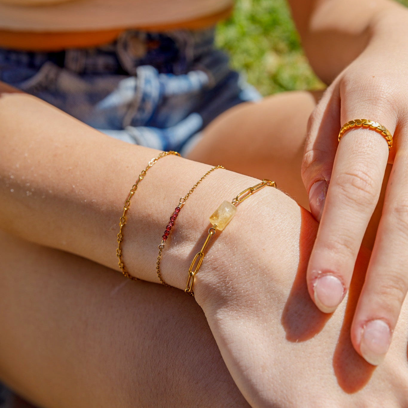 A close-up lifestyle shot of a person's wrist and hand wearing multiple delicate gold bracelets, including a textured chain bracelet, a thin chain with a small red stone, and a chain with a pale yellow stone, alongside a matching gold ring on the finger.
