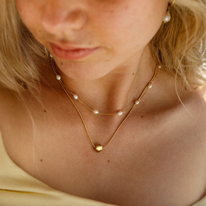 A close-up lifestyle shot of a blonde woman wearing a delicate double-layered necklace with small white pearls and gold chains, along with a wide-brimmed straw hat.