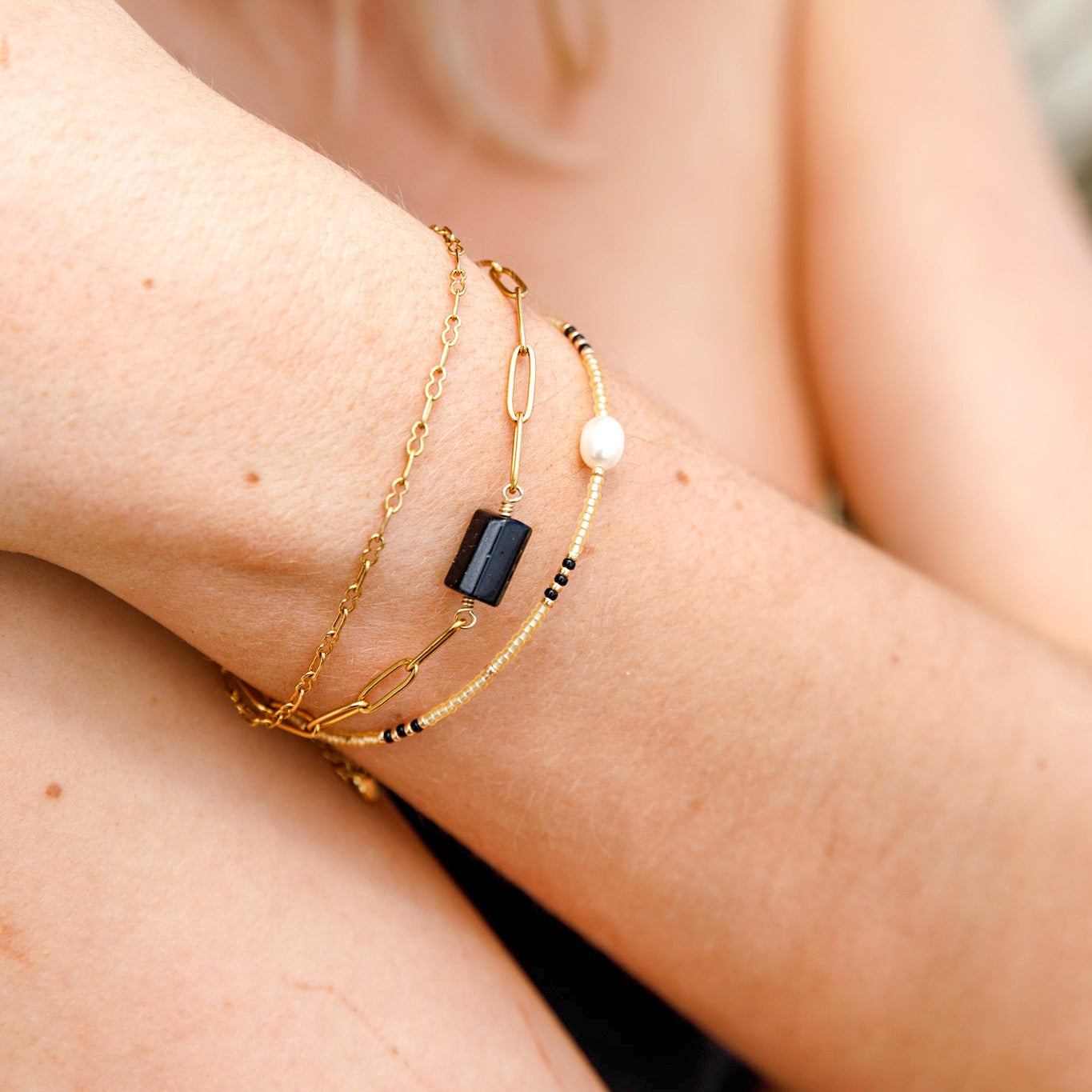 A close-up of a person's wrist wearing a delicate gold chain bracelet with a black rectangular stone and a small white pearl bead.