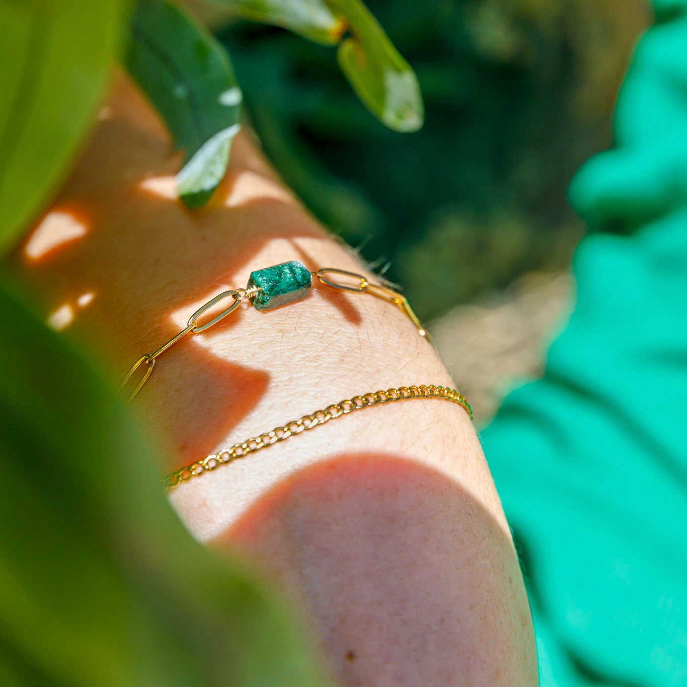 A close-up image of a gold-colored bracelet with a green gemstone on a person's wrist, surrounded by green leaves.