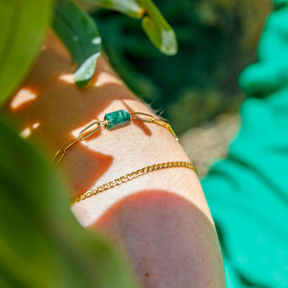 A close-up image of a gold-colored bracelet with a green gemstone on a person's wrist, surrounded by green leaves.