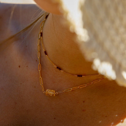 Close-up of a person's neck and upper chest wearing two layered gold necklaces, including one with a small rectangular clear gemstone pendant, under warm natural light.