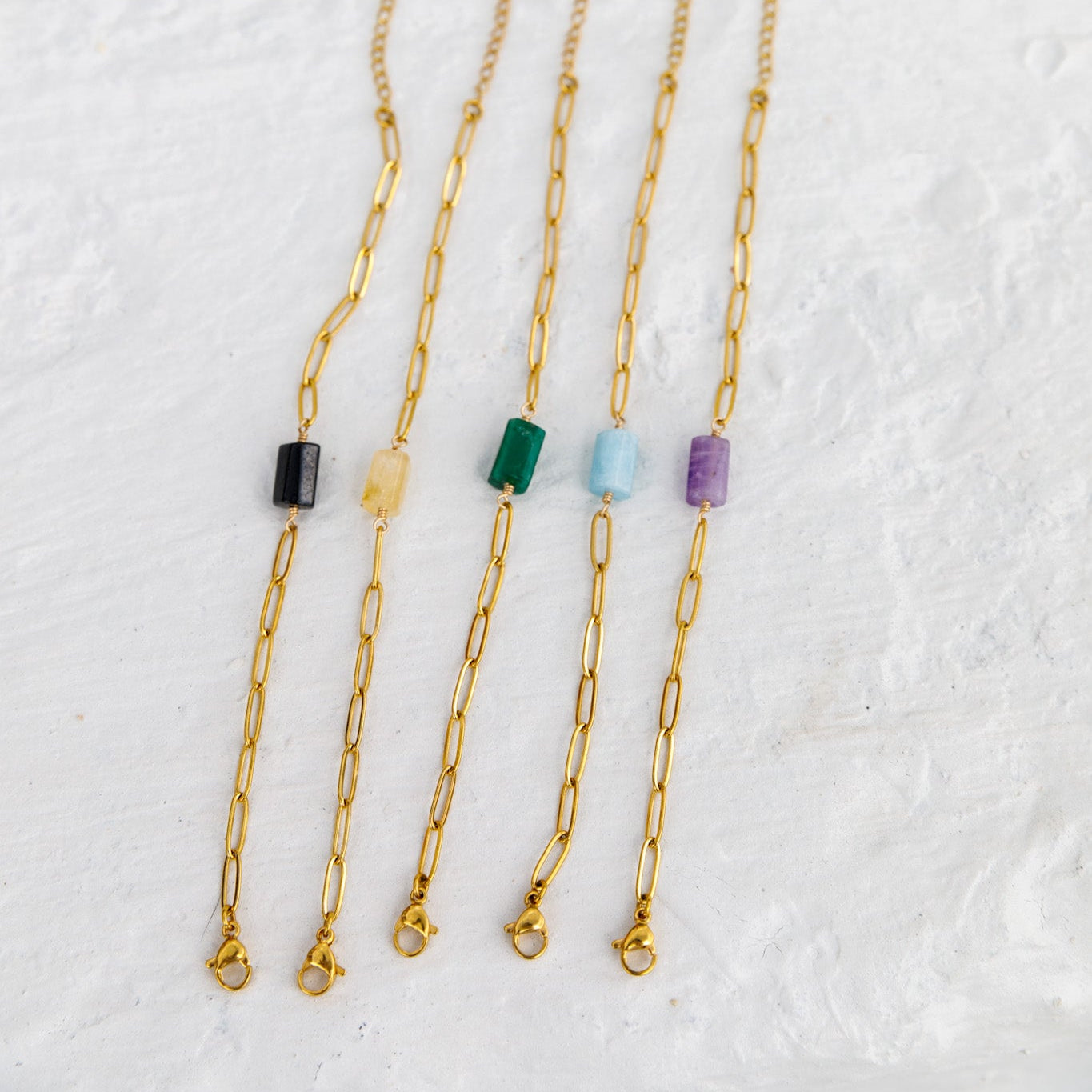 Four gold paperclip chain necklaces with small rectangular gemstone beads in black, yellow, green, and purple, arranged on a textured white background.