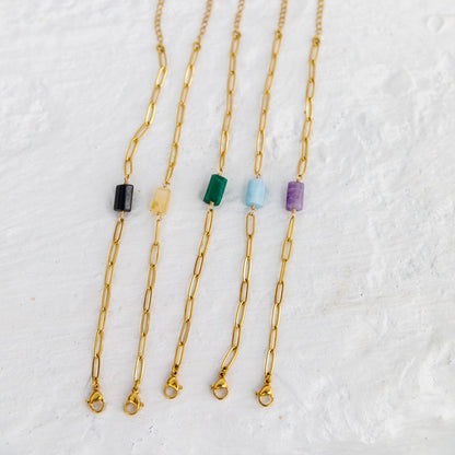 Four gold paperclip chain necklaces with small rectangular gemstone beads in black, yellow, green, and purple, arranged on a textured white background.