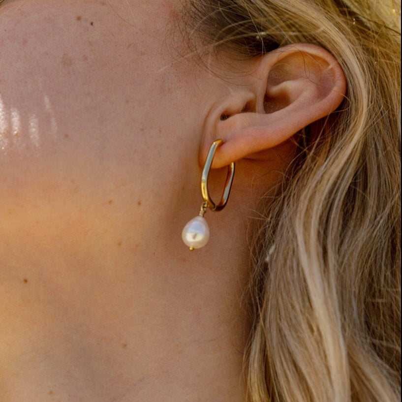 A close-up of a woman's ear wearing a gold huggie hoop earring with a dangling white freshwater pearl.