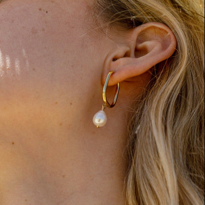 A close-up of a woman's ear wearing a gold huggie hoop earring with a dangling white freshwater pearl.