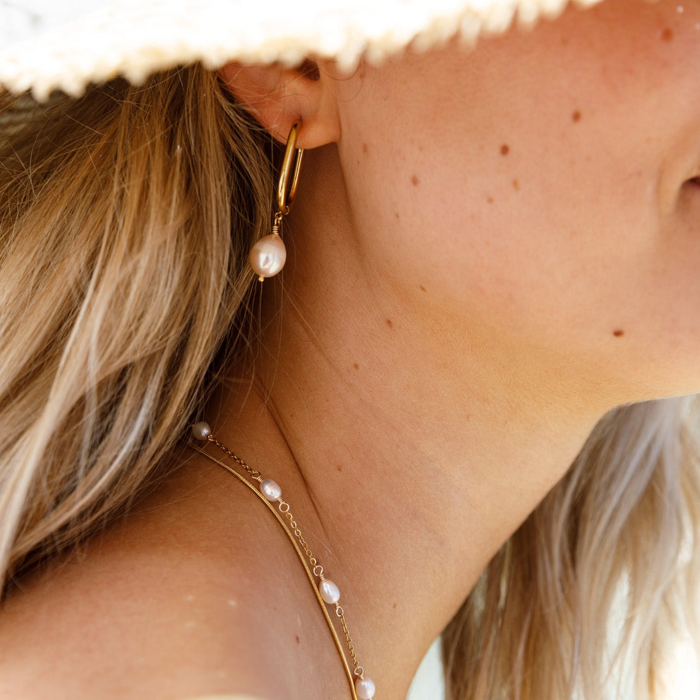 A close-up lifestyle shot of a woman wearing a gold hoop earring with a dangling white pearl, along with a delicate gold chain necklace with small pearls, under a white straw hat.