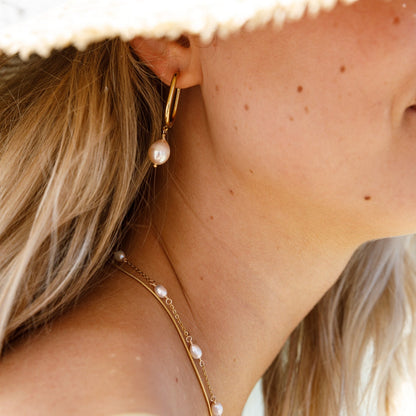 A close-up lifestyle shot of a woman wearing a gold hoop earring with a dangling white pearl, along with a delicate gold chain necklace with small pearls, under a white straw hat.